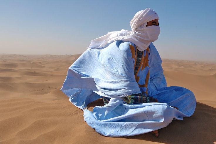 a man sitting on top of a sandy beach covered in a blue blanket and covering his face