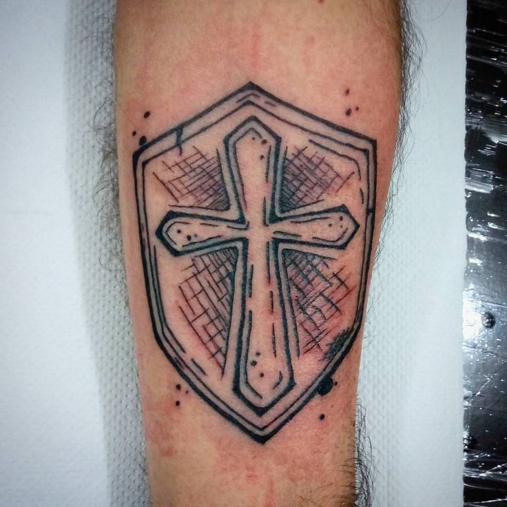 a cross tattoo on the leg of a man with a shield behind it that is black and grey