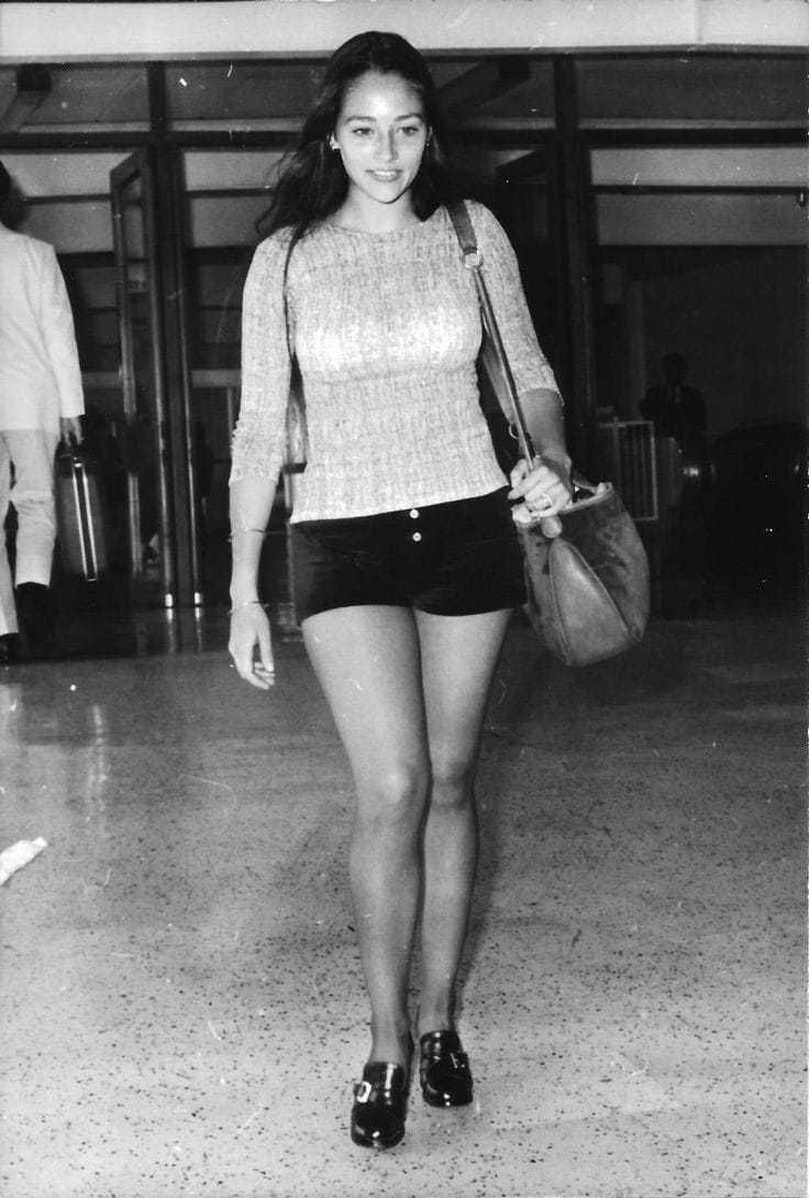 black and white photograph of a woman in short shorts walking through an airport with luggage