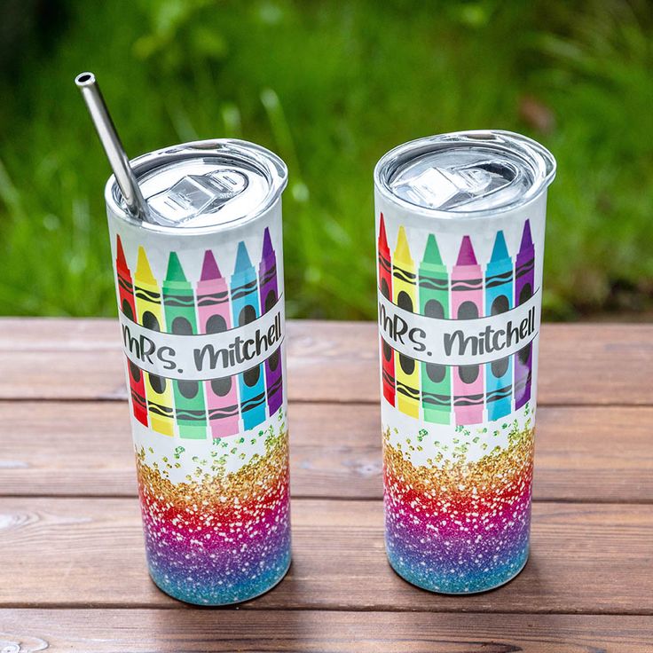 two colorful tumblers sitting next to each other on top of a wooden table with grass in the background