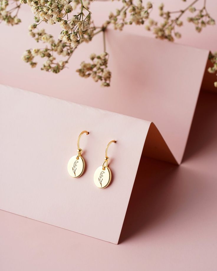 two small gold earrings sitting on top of a pink table next to some white flowers