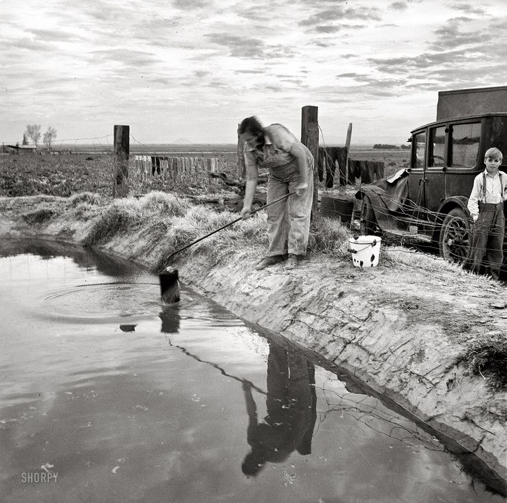 an old black and white photo of two people near a body of water with a truck in the background