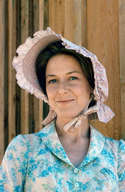 a woman wearing a blue shirt and a white hat smiles at the camera while standing in front of a wooden fence