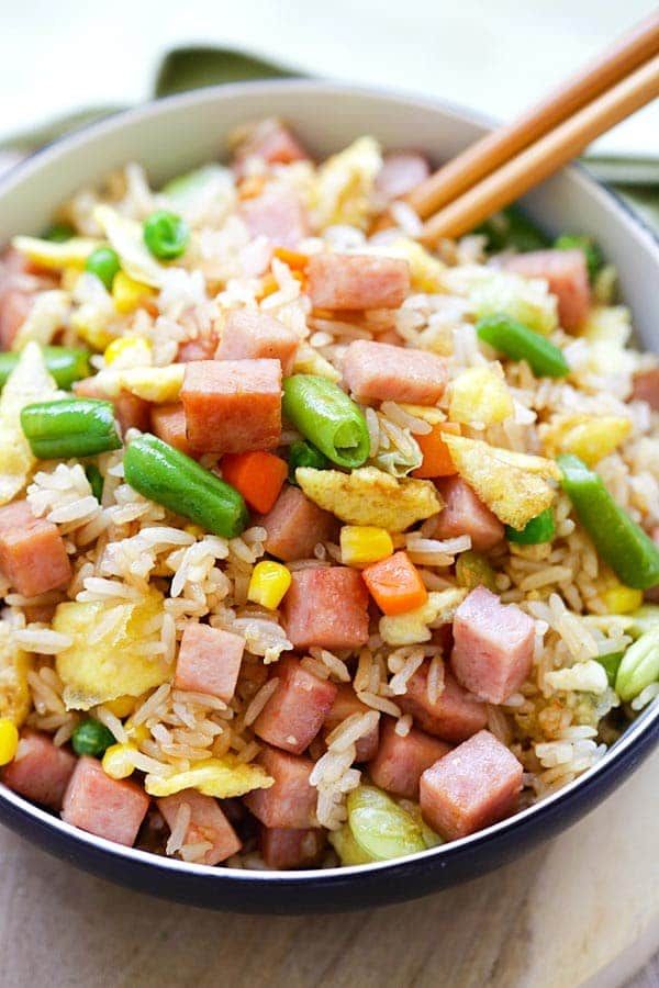 a bowl filled with rice, ham and vegetables next to chopsticks on a cutting board