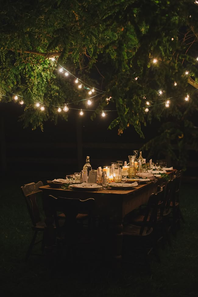 an outdoor dinner table is set with candles and wine bottles, surrounded by string lights