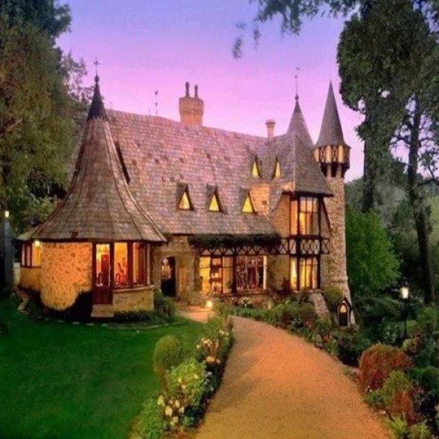 a large house with lots of windows and lights on it's front porch, surrounded by lush green trees