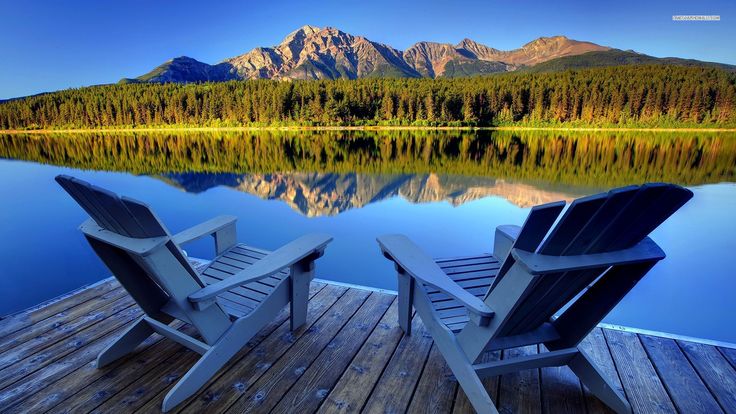 two adiron chairs on a dock overlooking a lake with mountains in the back ground