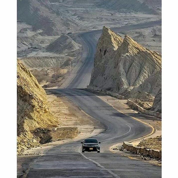 a car driving down the middle of a desert road in front of large rocks and mountains