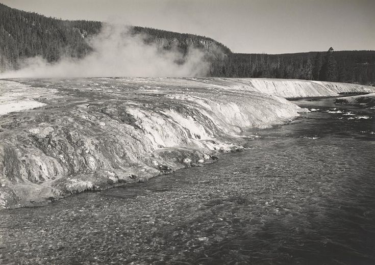 steam rises from the ground near a river in an area with snow covered mountains and trees