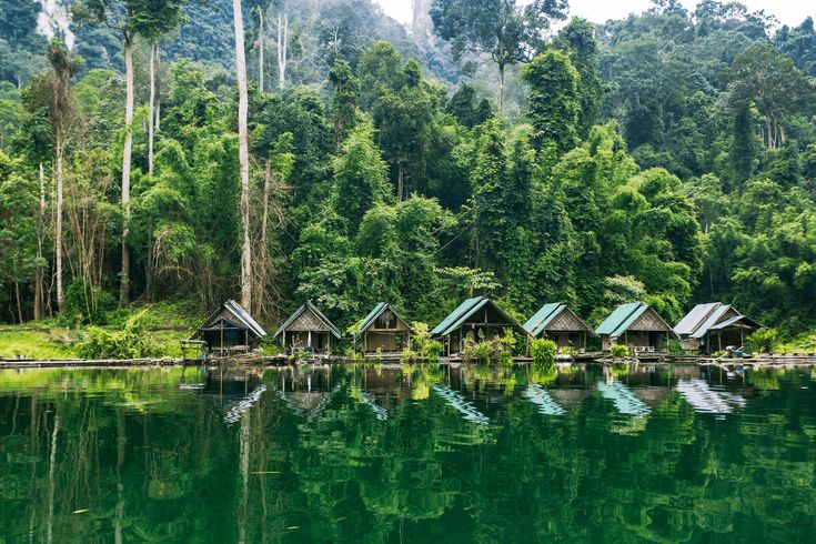 some huts are sitting on the shore of a lake in front of trees and mountains