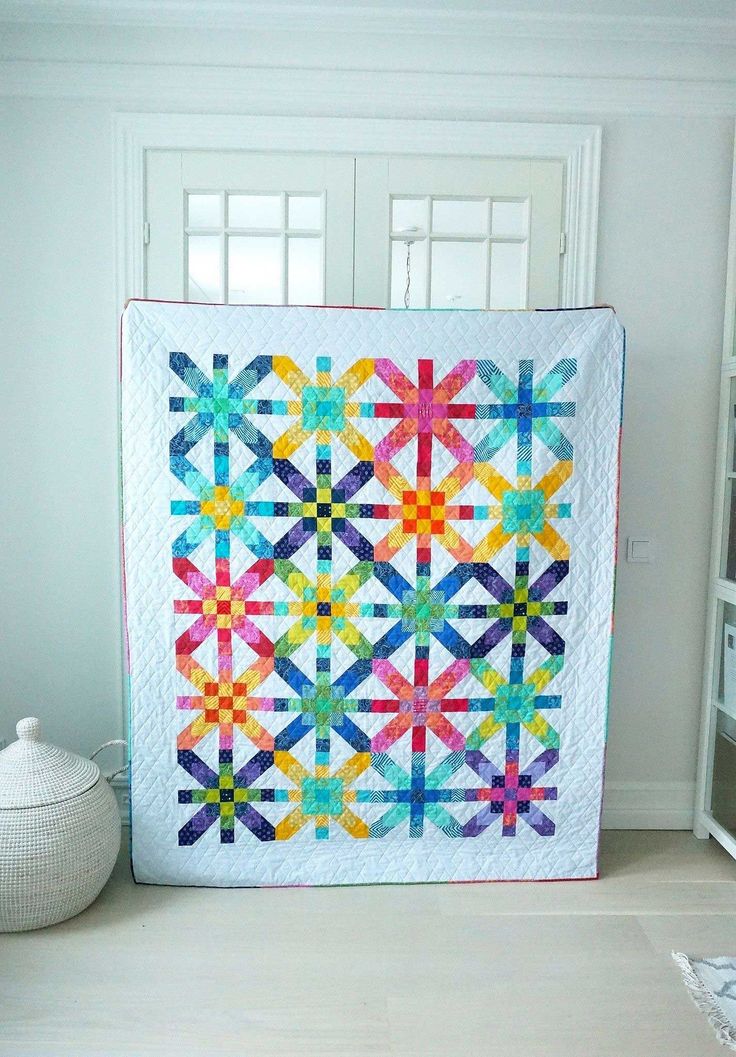 a colorful quilt hanging on the wall next to a white vase and window sill