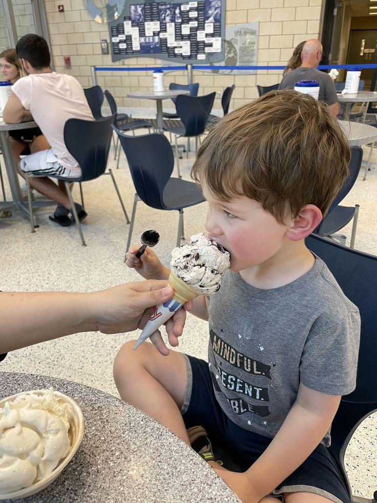 a young boy sitting at a table eating an ice cream cone with someone's hand