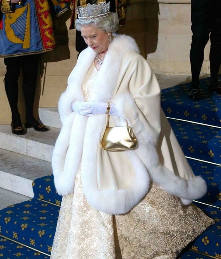 the queen of england is dressed in white fur and holding a gold purse as she walks up steps