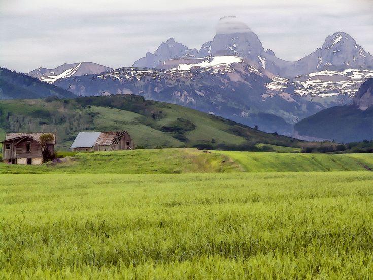 an old barn sits in the middle of a green field with mountains in the background