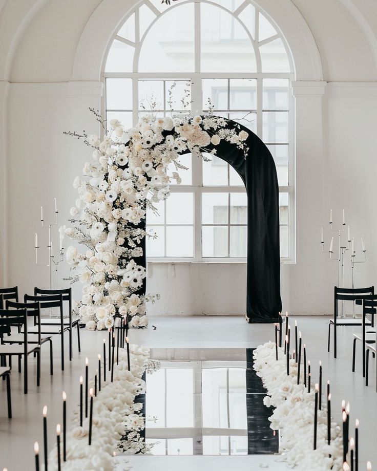 an arch decorated with flowers and candles stands in the middle of a white room filled with black chairs