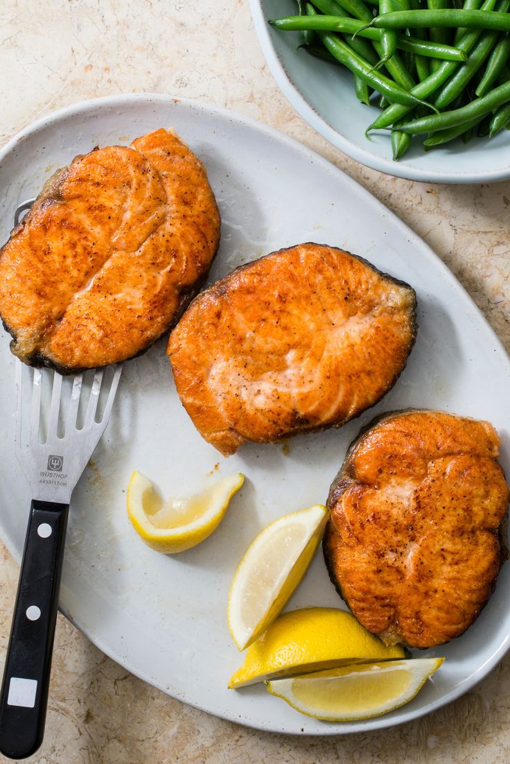 two pieces of fish on a plate with lemon wedges and green beans next to it