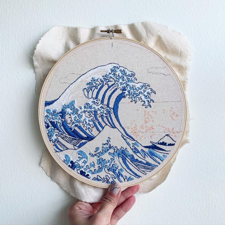 a hand holding up a blue and white embroidery art piece with the great wave on it