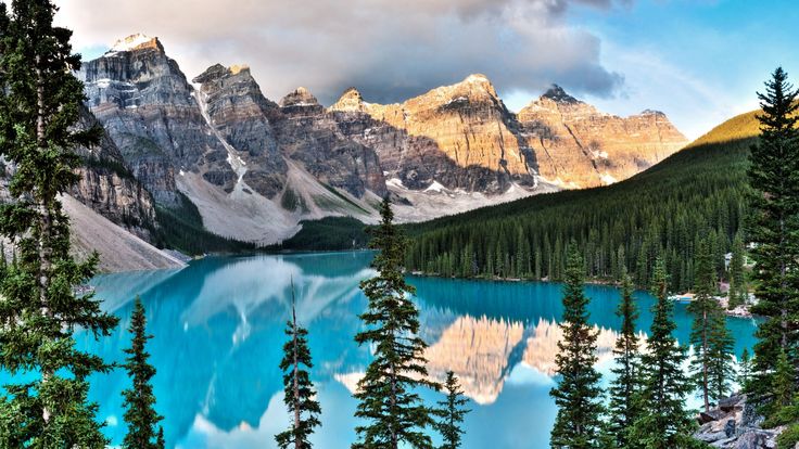 the mountains are covered in snow and blue water with pine trees around them, as well as clouds