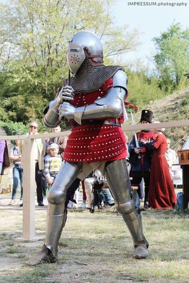 a man dressed in armor standing next to a wooden fence with other people around him