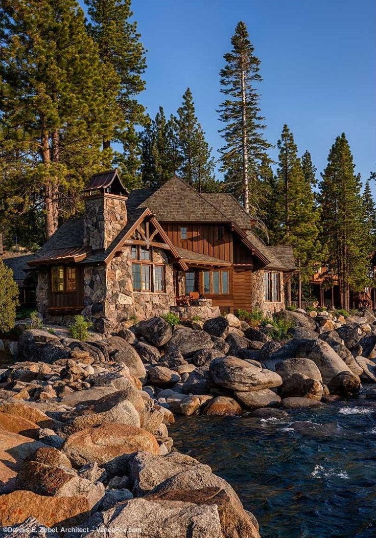 a log cabin sits on the shore of a lake surrounded by rocks and pine trees