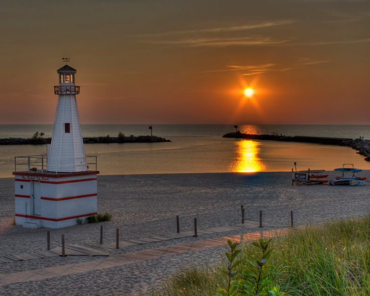 the sun is setting behind a light house on the beach with boats in the water