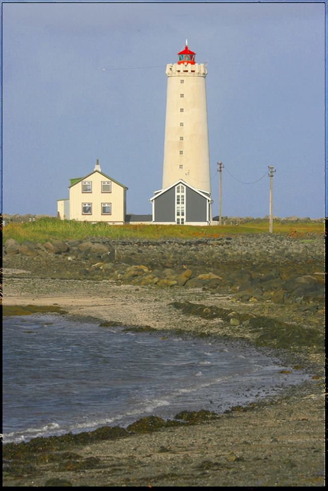 a lighthouse on top of a grassy hill next to the ocean with a building in the background