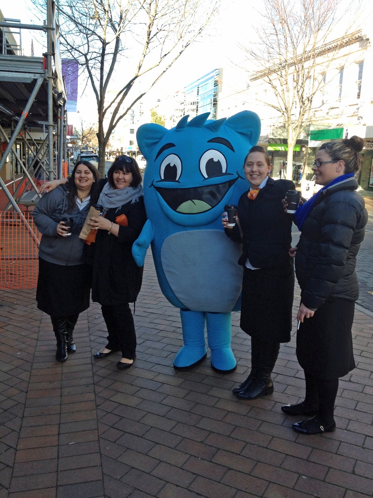 three women standing next to a blue monster mascot on a brick sidewalk with trees in the background