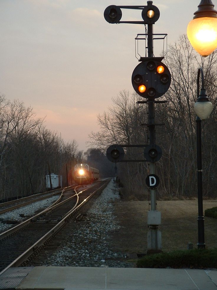 a train traveling down tracks next to a forest at dusk with the lights on and one light on