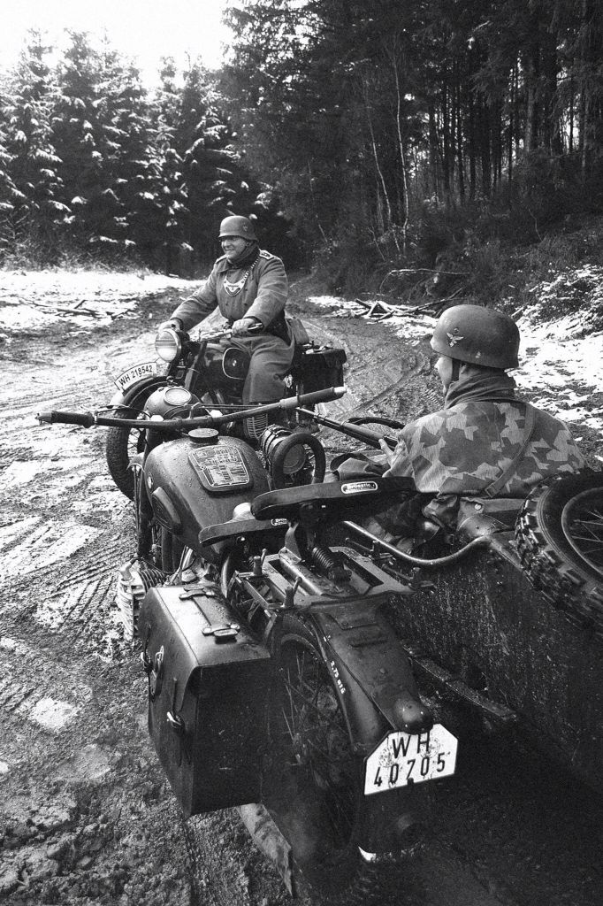 black and white photograph of two men riding on atvs in the snow with trees behind them