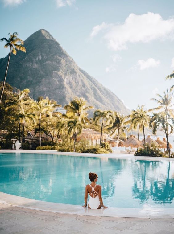 a woman sitting on the edge of a swimming pool in front of mountains and palm trees