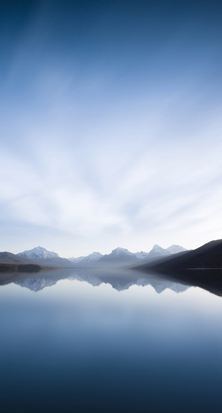 the sky is reflected in the still water and mountains are visible behind it, as seen from across the lake