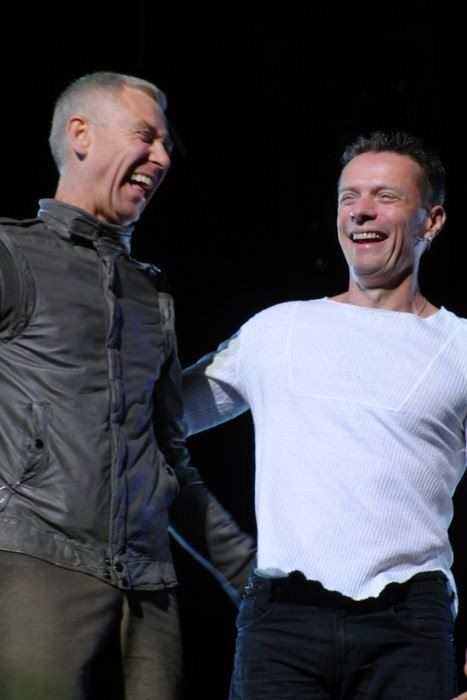 two men laughing and standing next to each other on stage with one holding his arm out