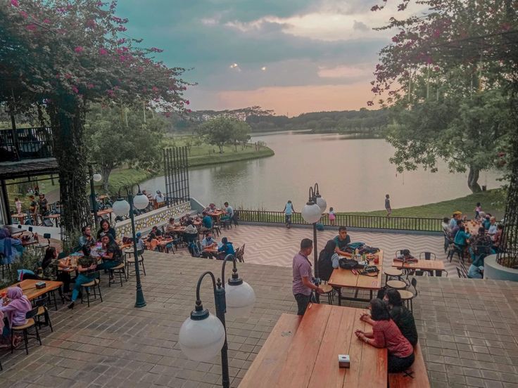 people are sitting at picnic tables in front of a lake with trees and benches around it