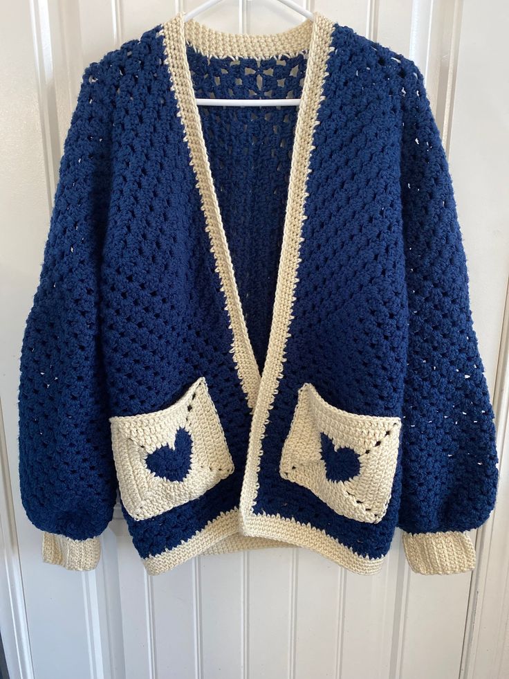 a crocheted blue jacket hanging on a white door hanger with an open front pocket