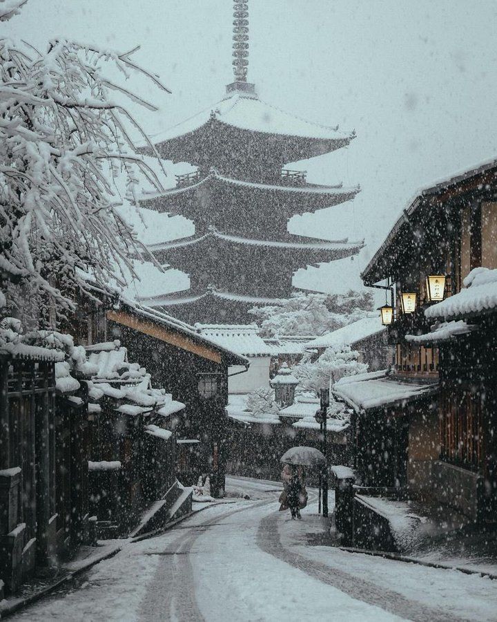 a snow covered street with buildings and lights in the distance, surrounded by snow falling on the ground