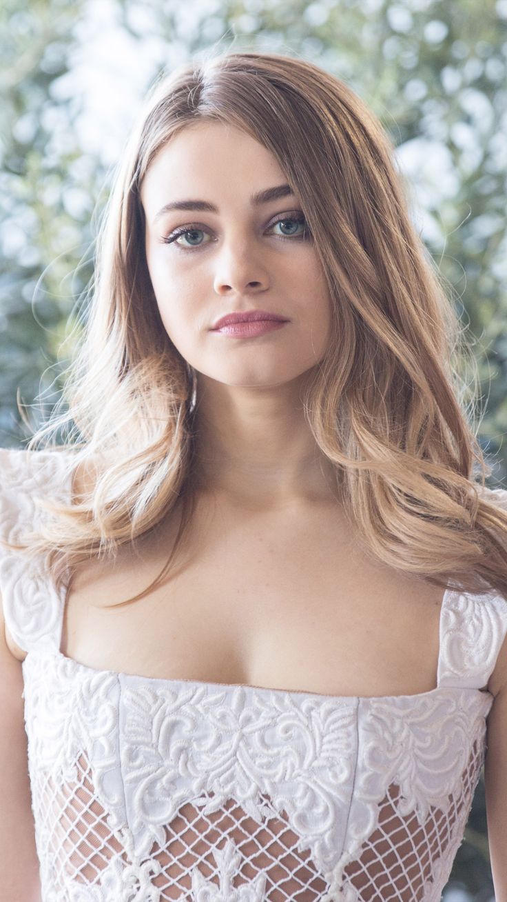 a woman with long hair wearing a white dress and looking at the camera while standing in front of some trees