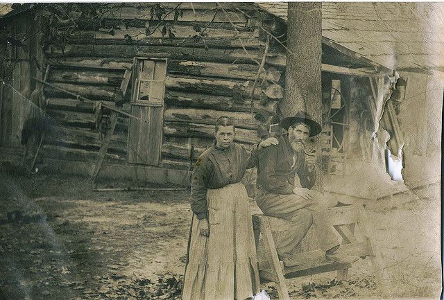 an old black and white photo of two men standing next to each other in front of a log cabin