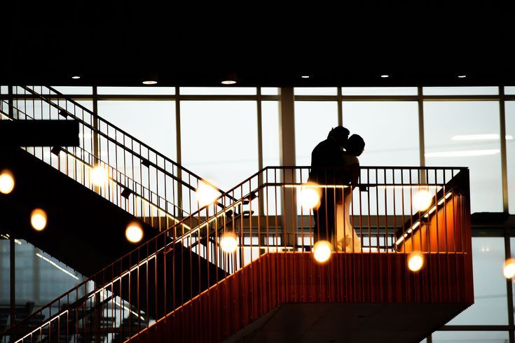 two people standing on top of a stair case next to each other in front of large windows