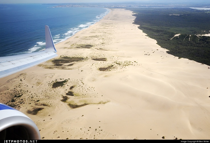 an airplane wing flying over a sandy beach next to the ocean