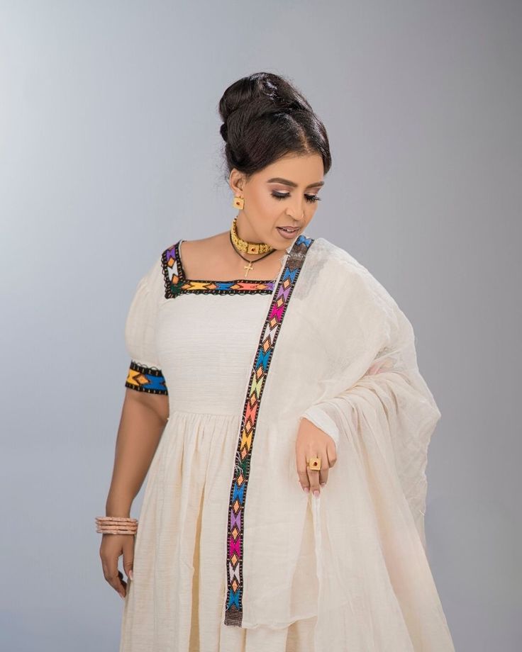 a woman wearing a white dress with colorful details on the neck and sleeves, standing in front of a gray background