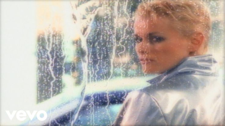 a woman with blonde hair standing in front of a rain soaked window and looking at the camera