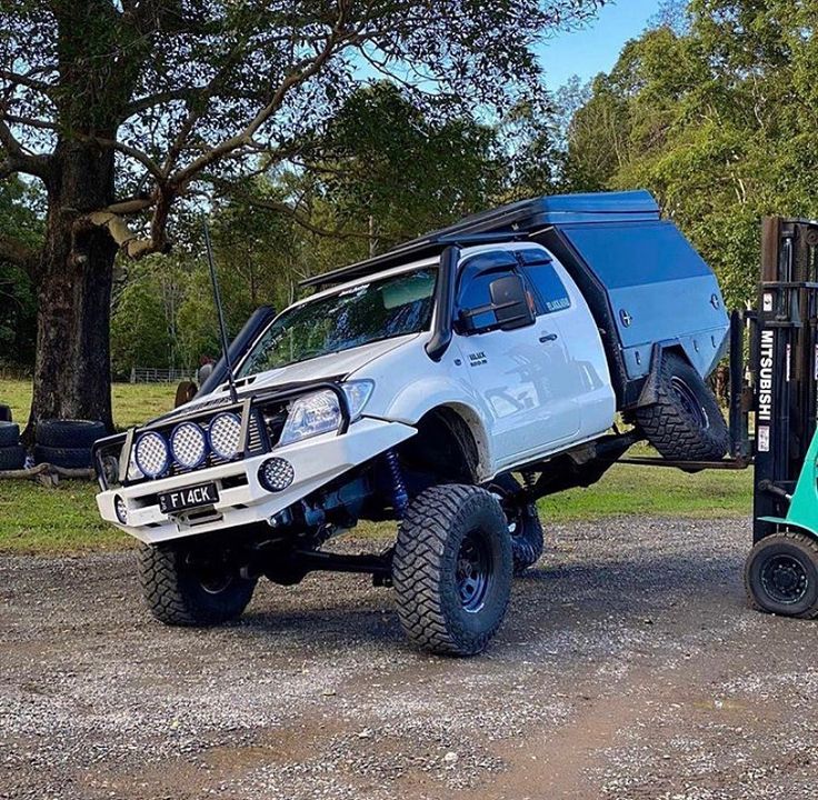 a white truck parked next to a forklift on top of a dirt road with trees in the background