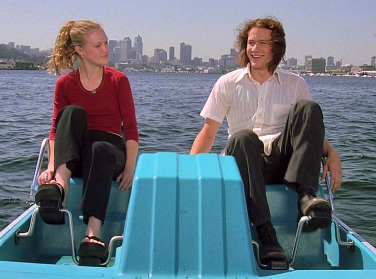 a man and woman sitting on the back of a blue boat in water with city skyline behind them