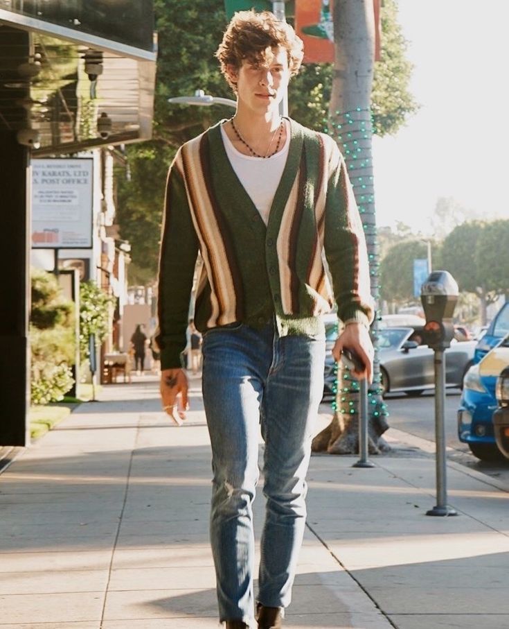 a young man is walking down the street wearing jeans and a striped cardigan sweater