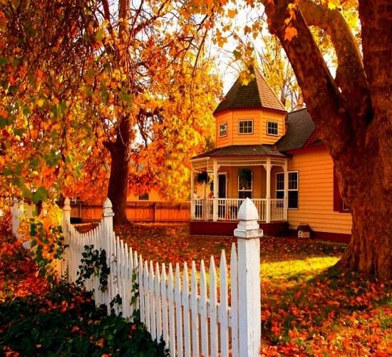 a white picket fence in front of a yellow house with fall leaves on the ground