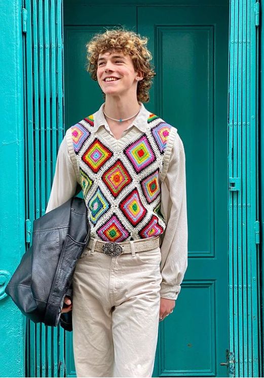 a young man standing in front of a blue door wearing a crochet sweater