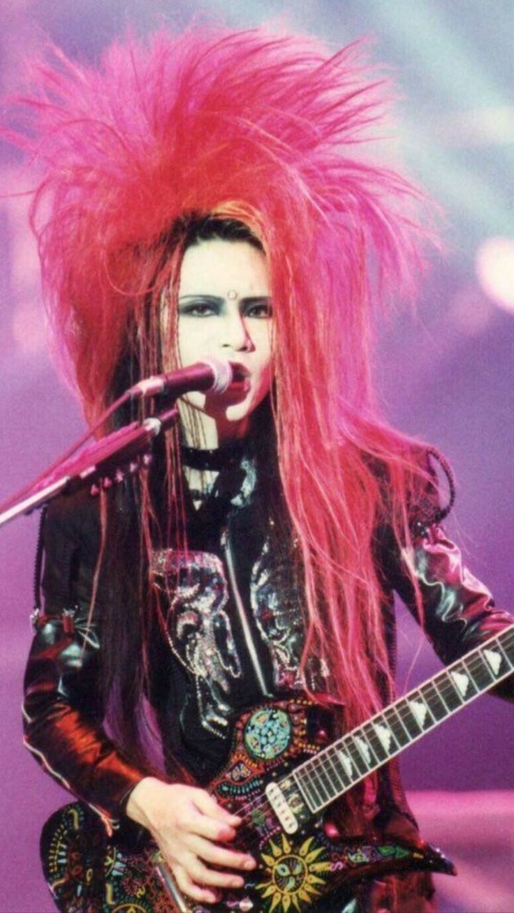 a woman with pink hair and makeup playing guitar in front of a microphone on stage