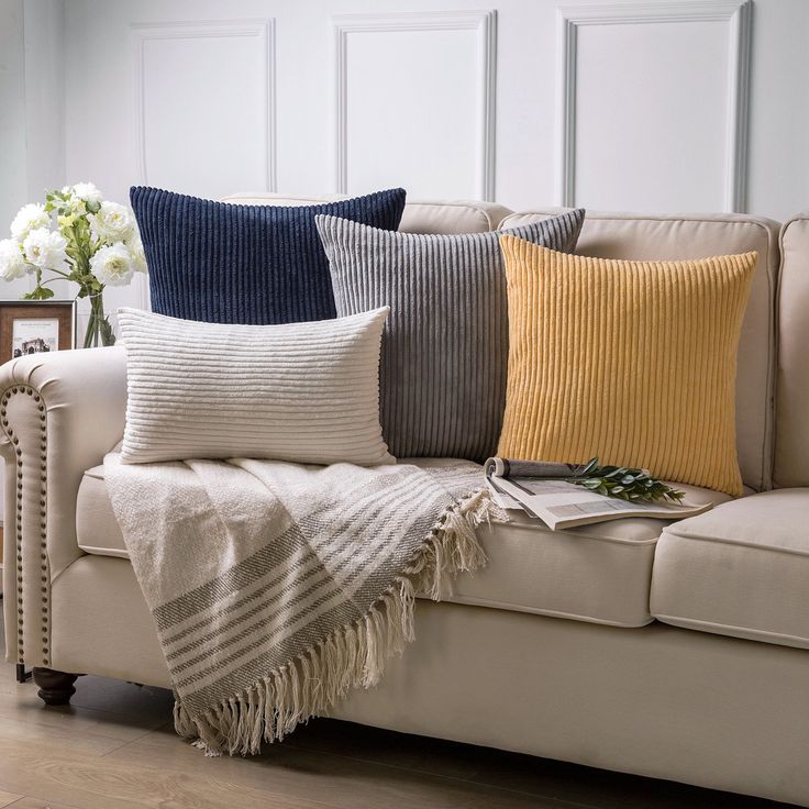 a couch with four different colored pillows on it and a vase full of flowers in the corner