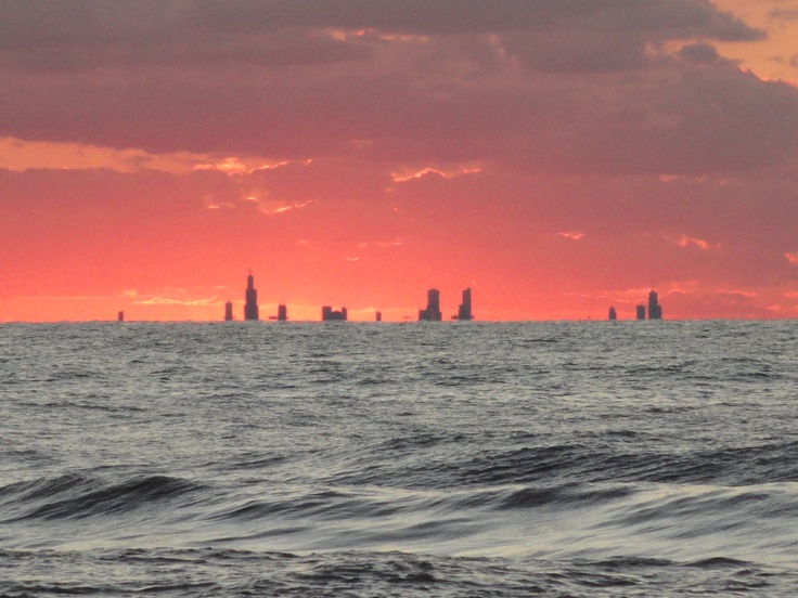 a large body of water with some buildings in the distance and clouds above it at sunset