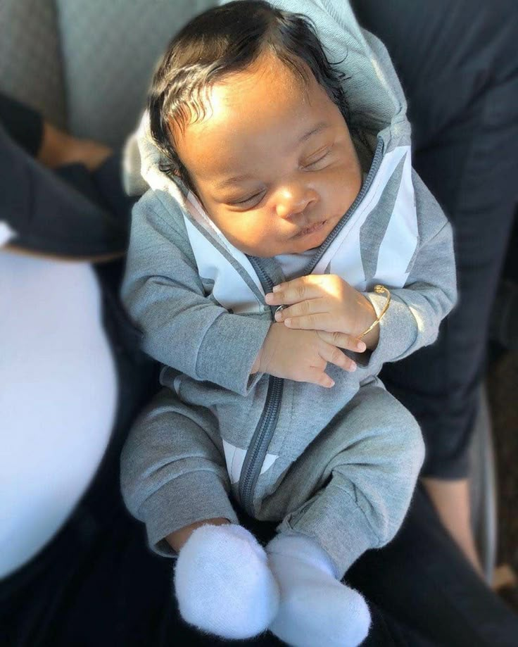 a baby sleeping in a car seat wearing a grey hoodie and white socks with his eyes closed